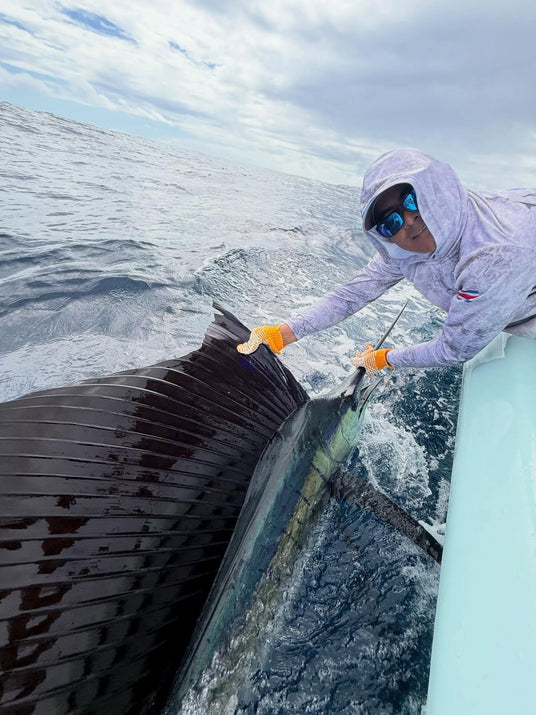 En sportfiskare i huva, solglasögon och handskar håller en stor segelfisk vid fenan över relingen på havet under molnig himmel.