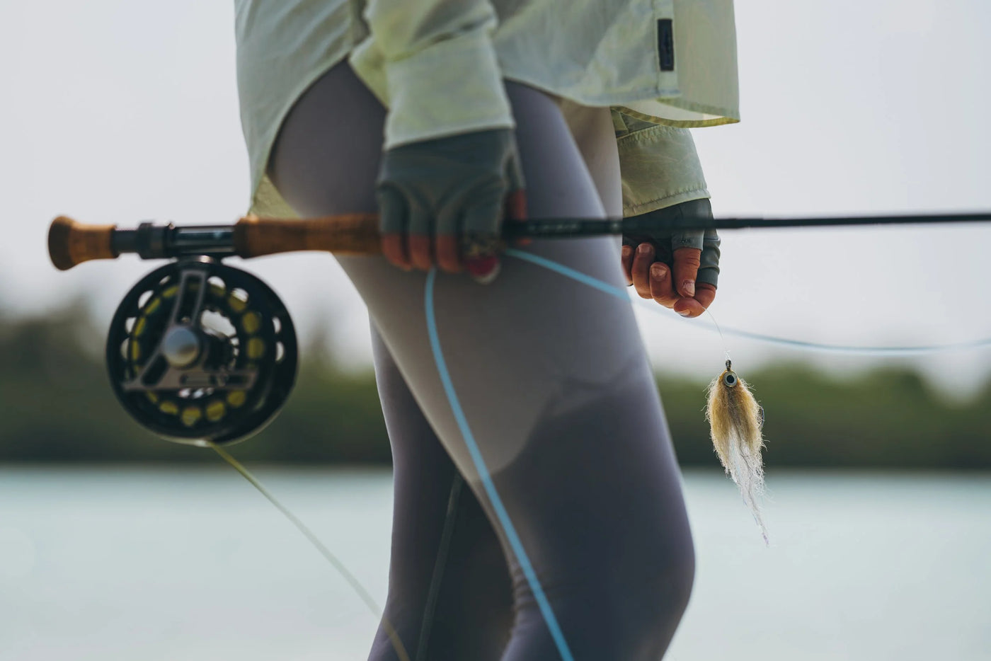 Sportfiskare i lätt klädsel med handskar håller ett flugfiskespö vid vattnet, redo för spännande fiskeäventyr.