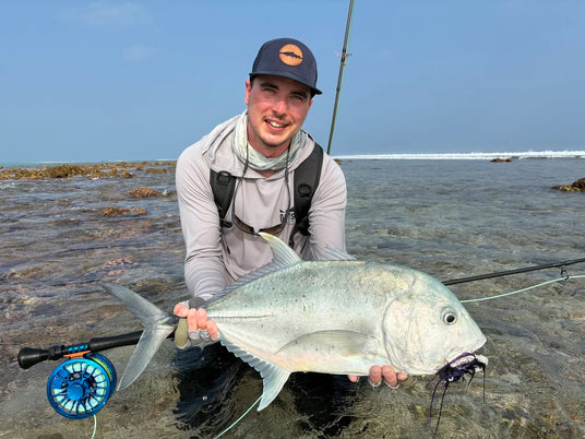 En sportfiskare knäböjer i grunt vatten med fiskespö och stor silverfisk, ler mot kameran. Hav och himmel syns i bakgrunden.