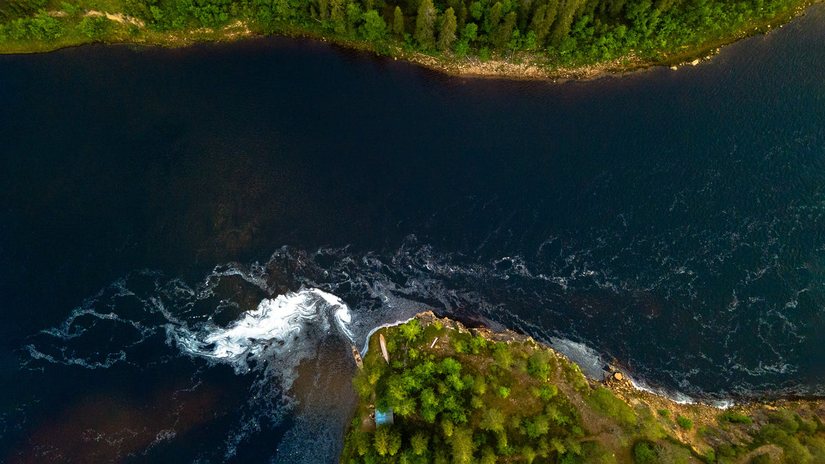 Flygfoto över en älv omgiven av tät grön skog, med virvlande vitt vatten vid stranden - perfekt för sportfiskare på fiskeresa.