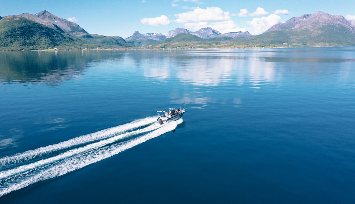 En motorbåt kör snabbt över en blå sjö med vita svallvågor. Gröna berg och klar himmel - idealiskt för sportfiskeresa.