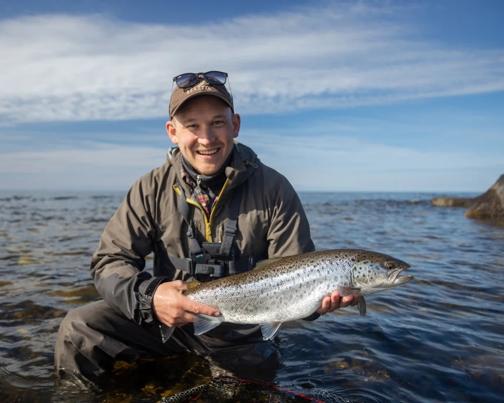 Sportfiskare i utrustning håller en stor fisk, står i grunt vatten under blå himmel - glädje från en lyckad fiskeresa.