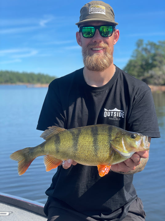 Skäggig man med solglasögon och hatt håller stor abborre på båt under fiskeresa, lugnt vatten och träd i bakgrunden.