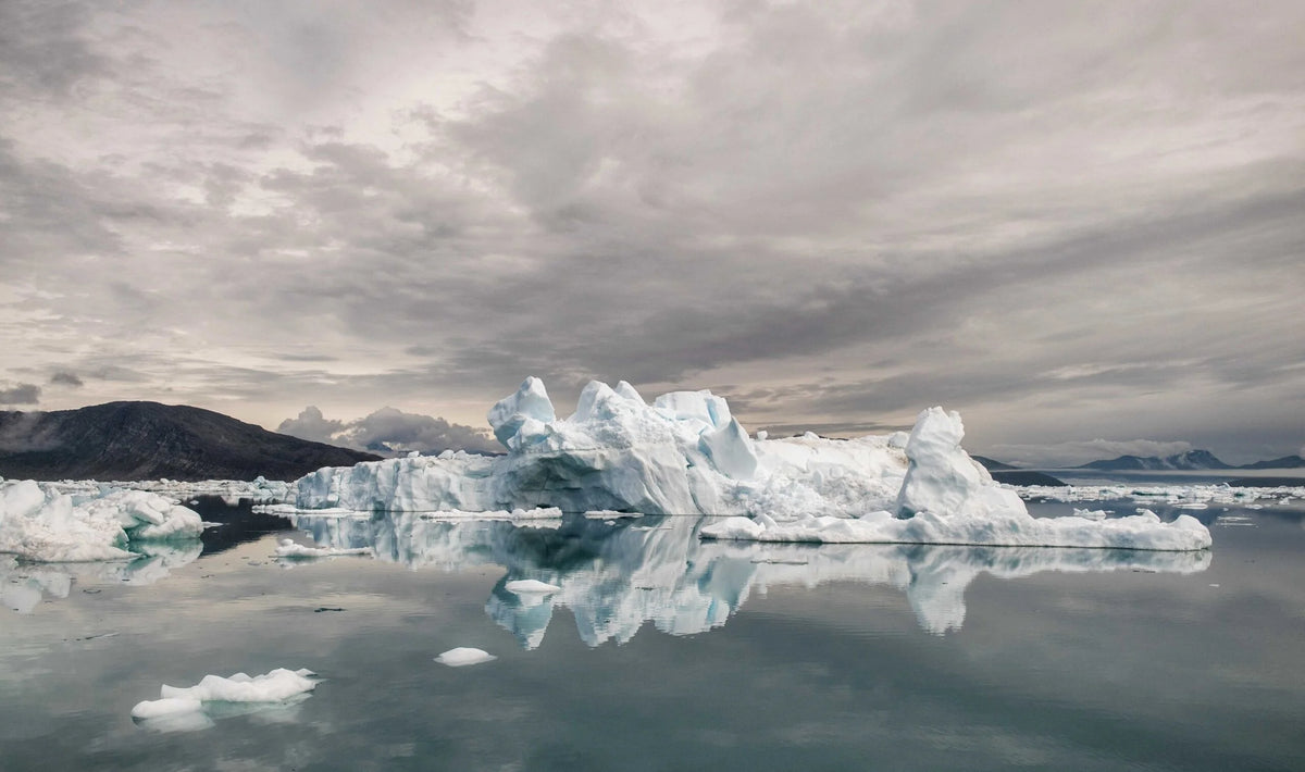 Ett stort isberg flyter på lugnt vatten under molnig himmel, med berg i fjärran - en inspirerande vy för din arktiska fiskeresa.