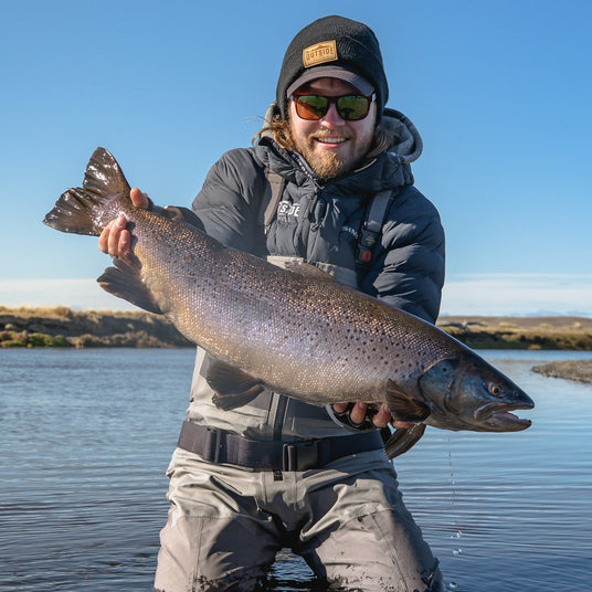 Leende sportfiskare i fiskeutrustning och solglasögon håller en stor fisk i grunt vatten under klar himmel på minnesvärd fiskeresa.