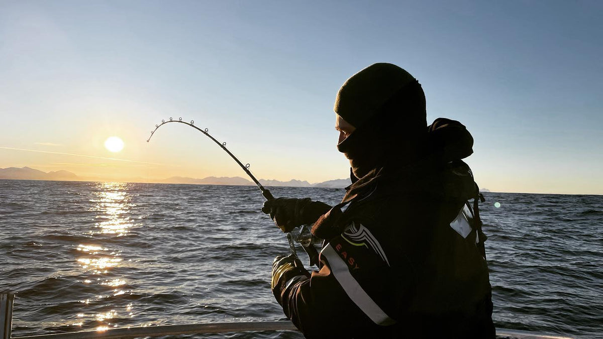 Sportfiskare på båt fiskar med böjt spö över lugnt hav vid gryning eller skymning, berg anas vid horisonten.