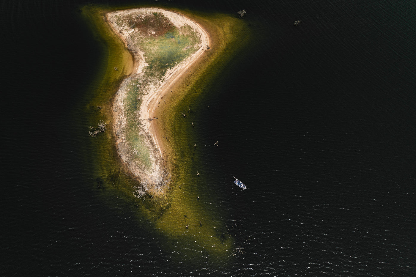 Flygfoto av liten, böjd ö med sandstrand och gräs, omgiven av mörkt vatten. Båt ankrad nära sportfiskare på land och i vattnet.
