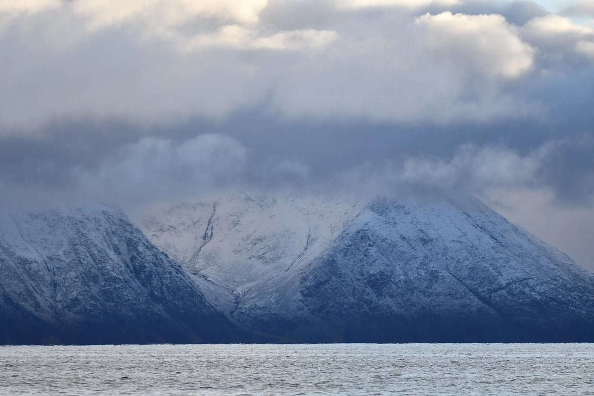 Snötäckta berg delvis dolda av moln reser sig bakom lugnt vatten under molnig himmel - perfekt för fiske och fiskeresa.