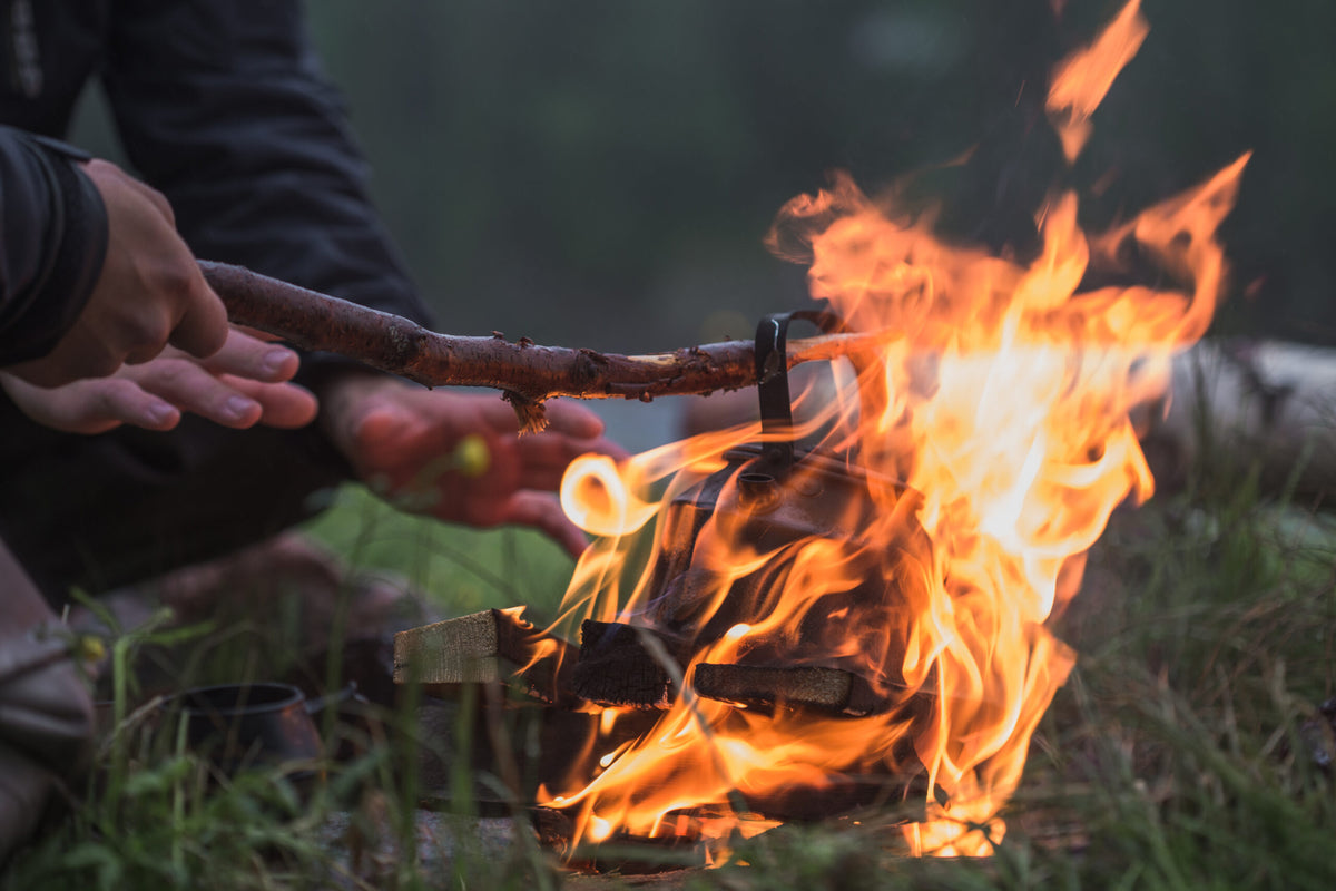 En person på fiskeresa grillar mat över öppen eld med pinne, lågorna stiger kring vedträn och gräs i förgrunden.