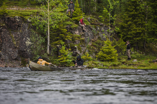 Två personer paddlar kanot på en flod, tre sportfiskare vid stenig skogskant bland gröna träd - perfekt för en fiskeresa.
