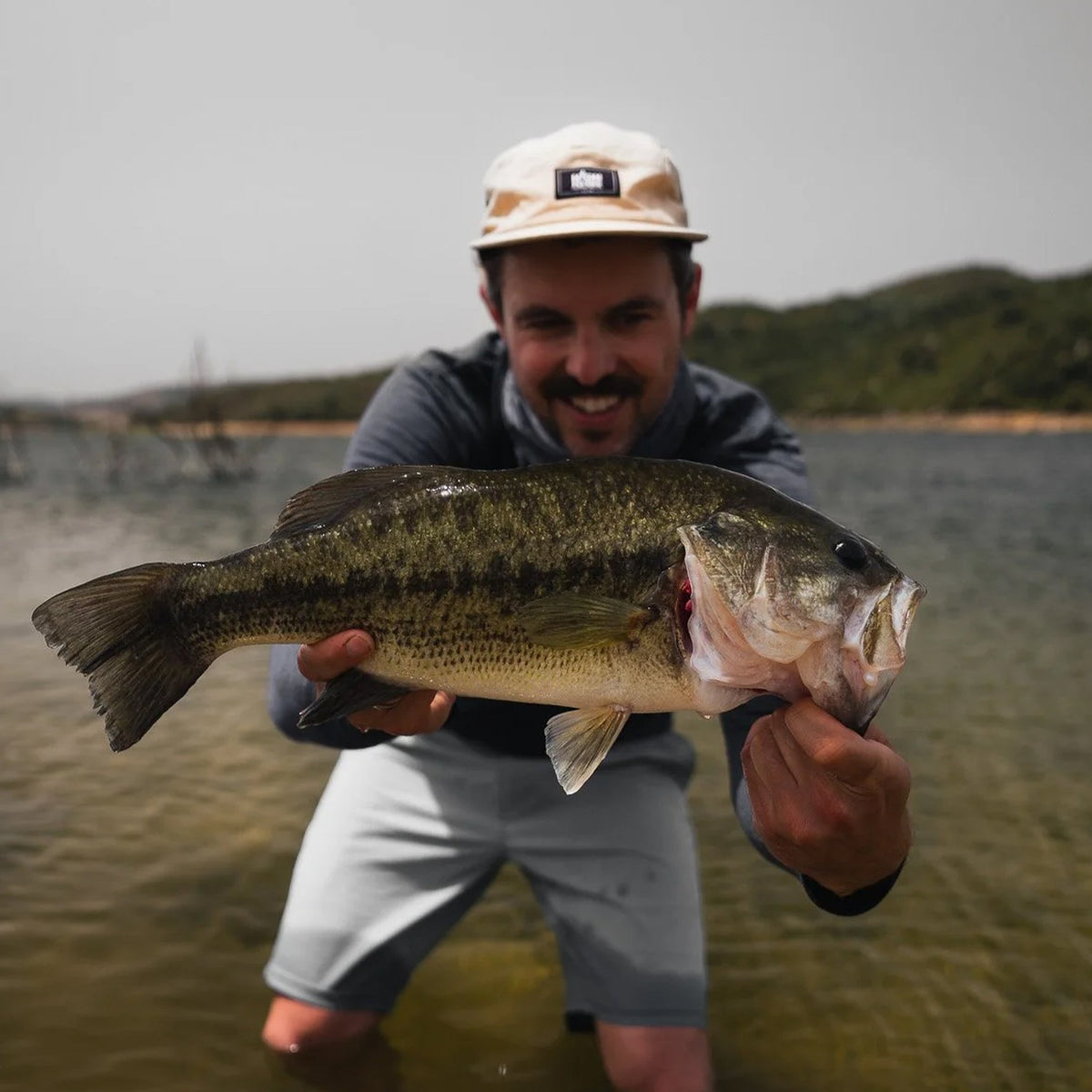 Glad man i hatt håller en black bass vid sjön på Sardinien med gröna kullar bakom