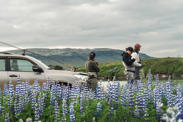 Tre personer står vid en bil med fiskespön, redo för en fisketur vid en flod. I bakgrunden syns berg och vilda blommor under en molnig himmel.