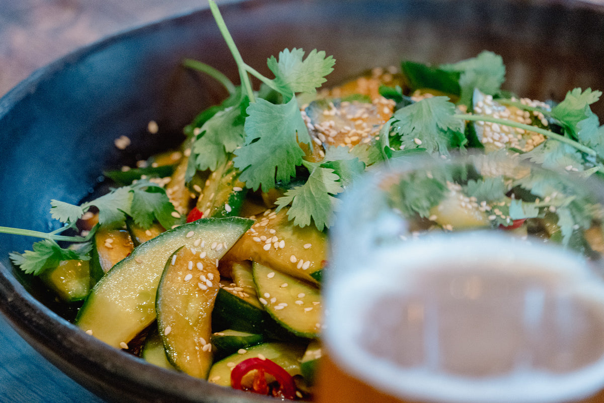 Närbild på gurksallad med koriander, sesamfrön och röd chili i en mörk skål – en perfekt avslutning på en fisketur, med en suddig öl i förgrunden.