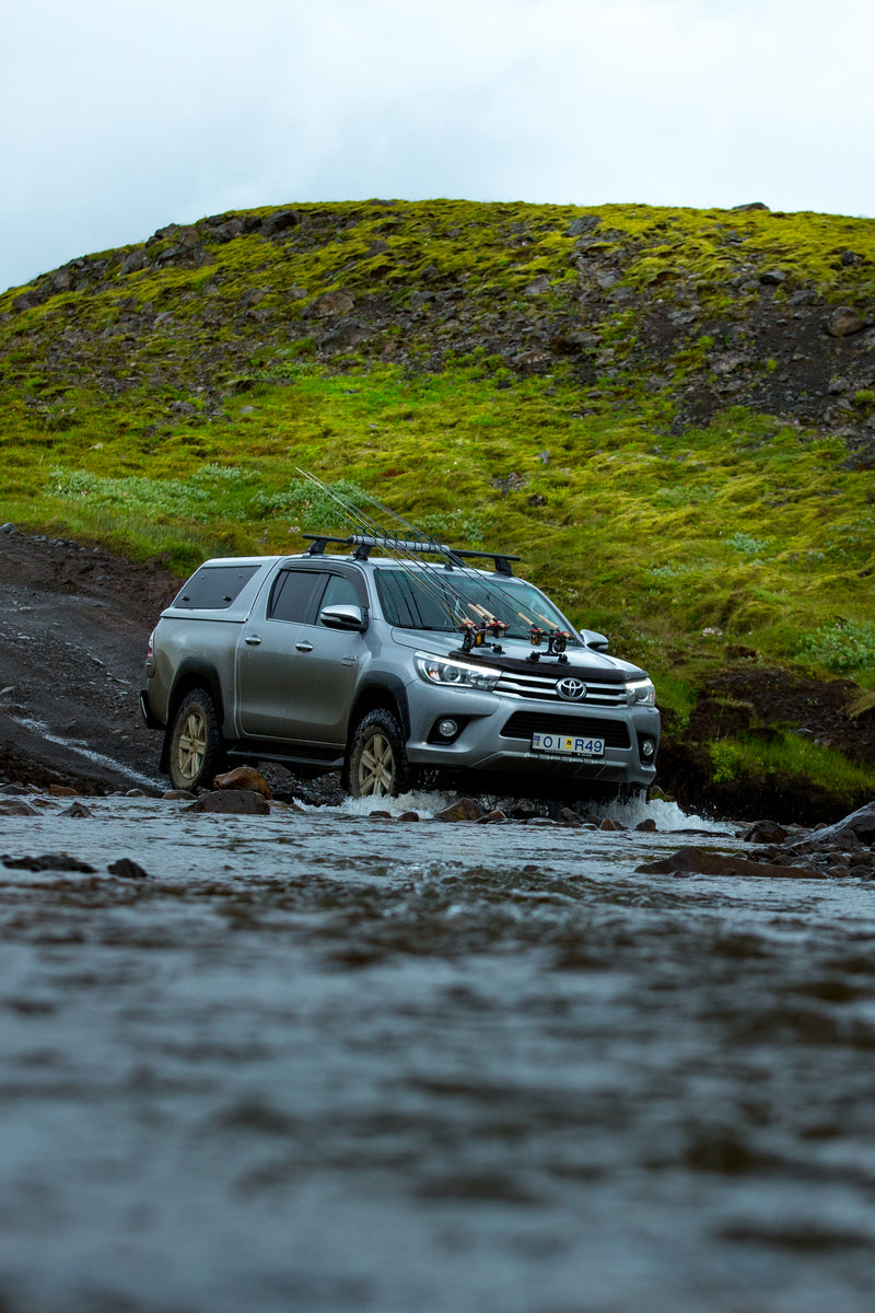 En silverfärgad Toyota-pickup kör genom en grund bäck på en lerig väg mot en grön kulle – perfekt för fiskeresor och fiskeäventyr.