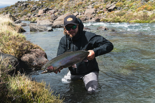 Sportfiskare med solglasögon och jacka knäböjer i en grund älv, håller en stor fångad fisk. Stenigt landskap och strömmande vatten.
