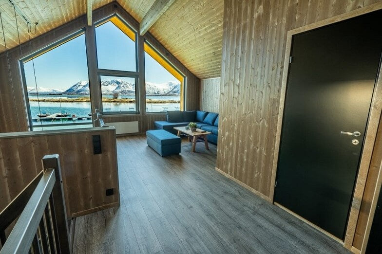 Träinteriör med stora fönster, utsikt över fjäll och vatten. Blå soffa, litet bord - perfekt för sportfiskarens avkoppling.
