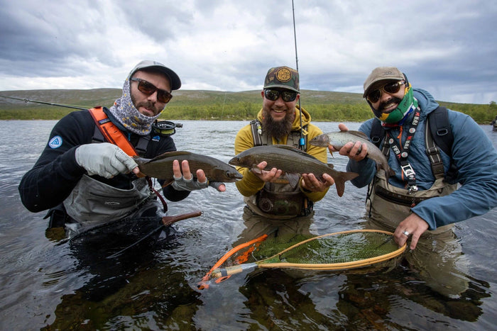 Tre leende sportfiskare i fiskekläder står knädjupt i älven med nyfångad fisk, håvar och spön under molnig himmel.