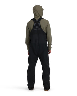 Person med ryggen mot kameran i khaki hoodie och svarta Simms GORE-TEX Pro fiskeoveraller, redo för fiskeresan.