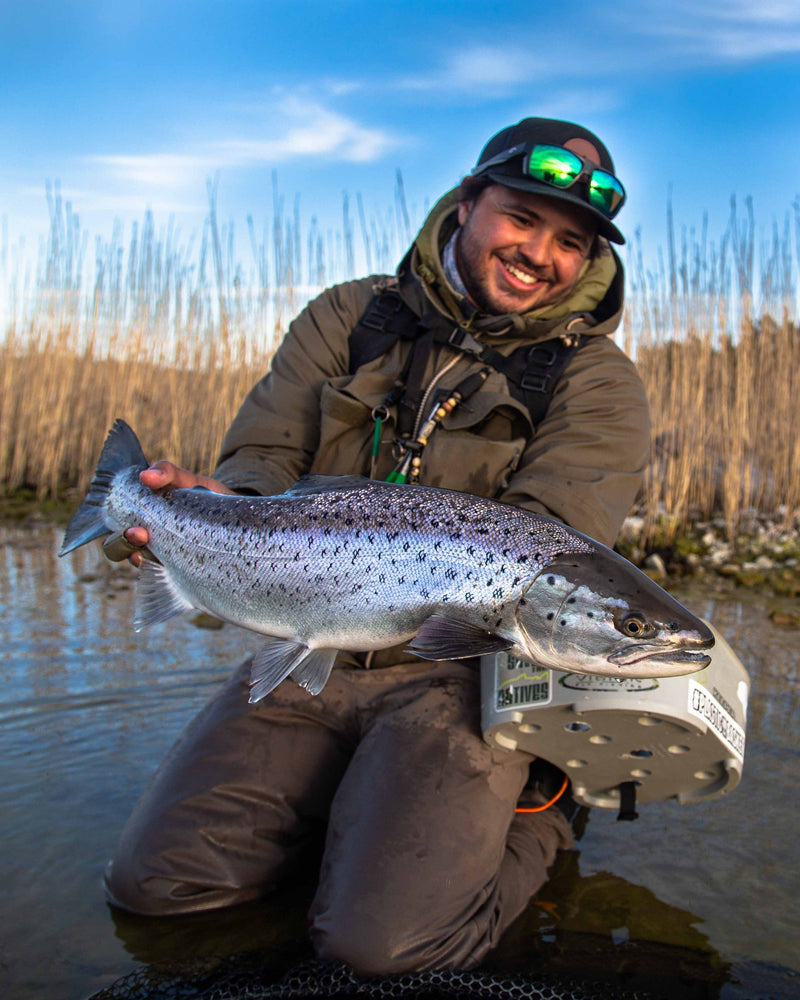 Leende sportfiskare i outdoor-kläder håller en stor, silverglänsande fisk vid vassen under blå himmel på lyckad fiskeresa.